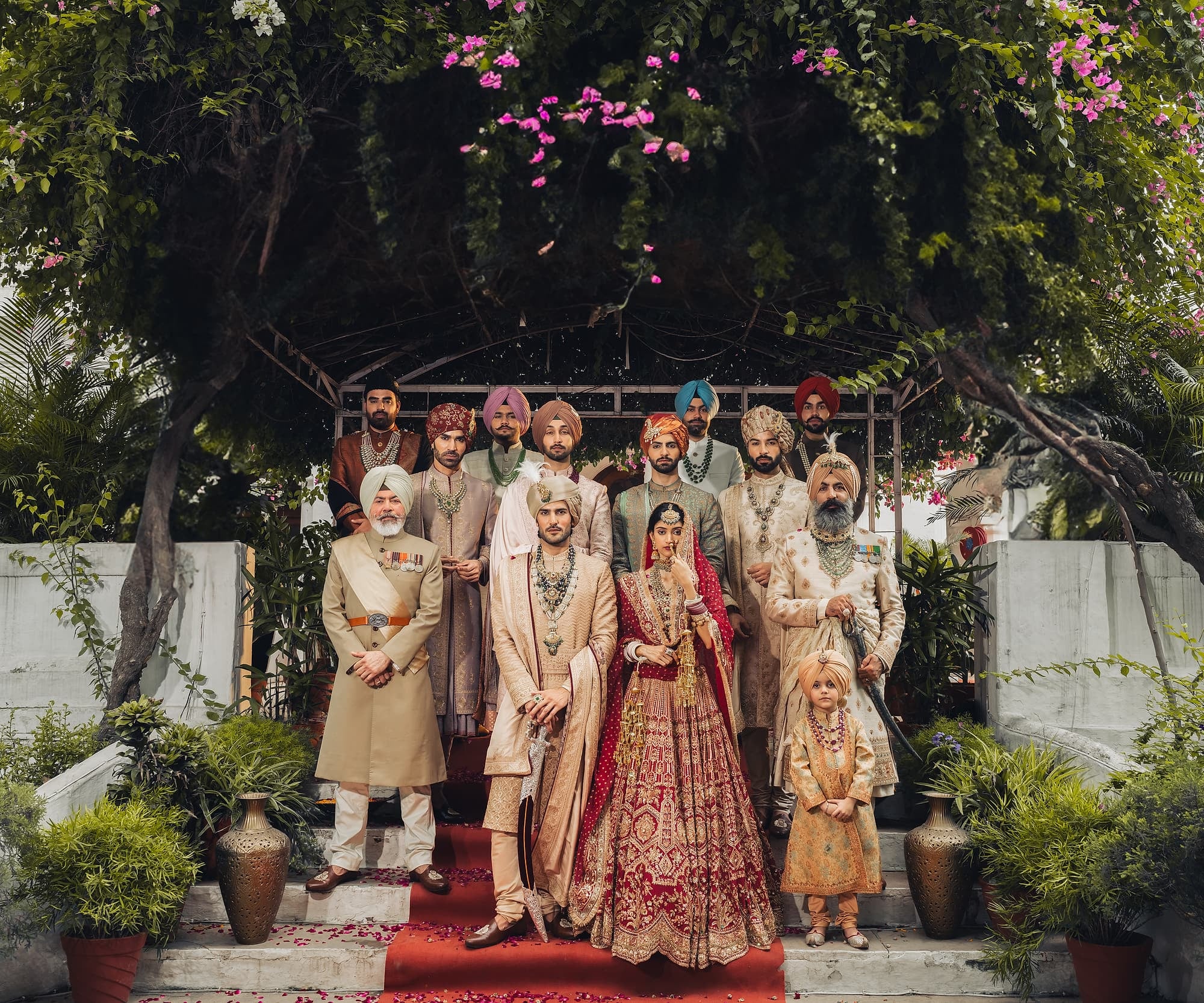 A wedding party assembled on the steps of a Panjabi haveli beneath a bougainvillea arbour — the groom in an ivory sherwani beside the bride in crimson, flanked by turbaned family